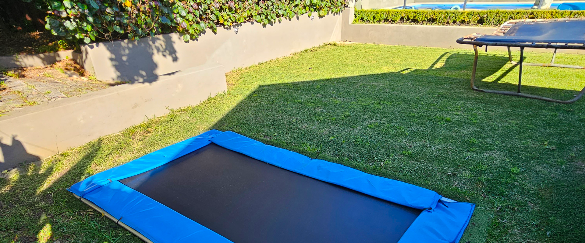 Rectangular inground trampoline on grass with a garden and pool area in the background